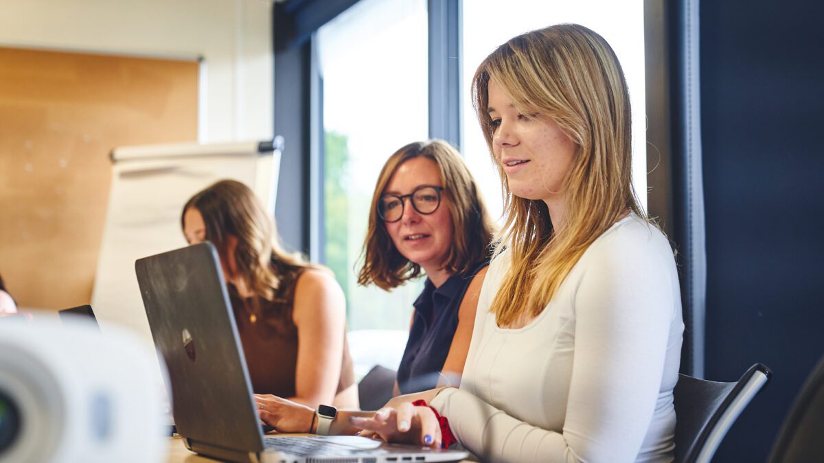 Three women working in an office, two are looking at a laptop screen together.