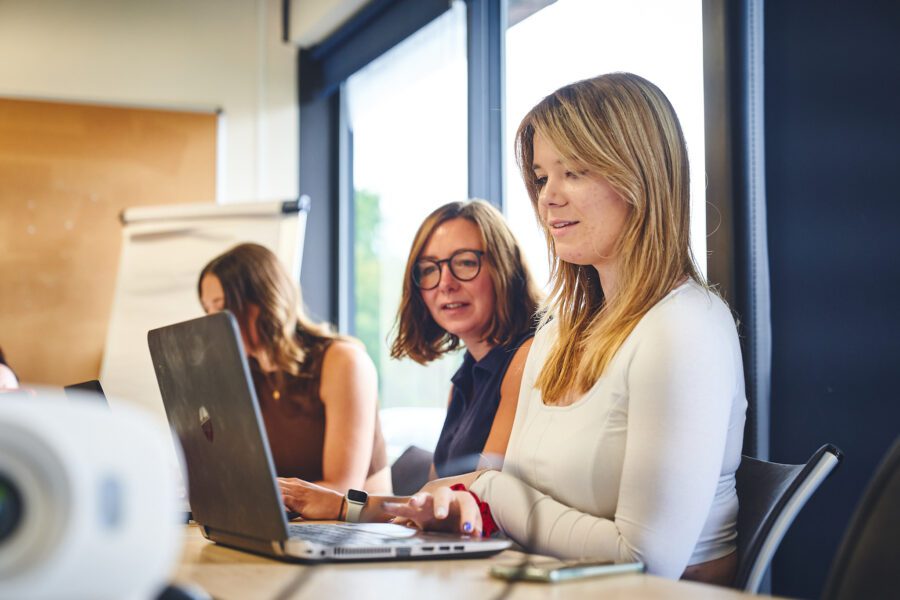 Three women working in an office, two are looking at a laptop screen together.