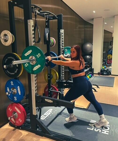 A woman in a sports bra and leggings lunging, leaning on a bar bell in a squat rack