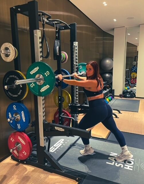 A woman in a sports bra and leggings lunging, leaning on a bar bell in a squat rack