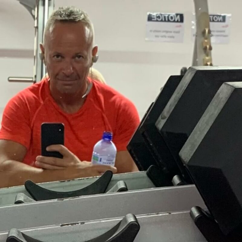 Mark leaning on gym equipment wearing an orange t-shirt