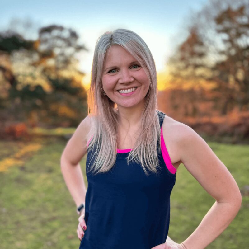 A woman standing in a garden with her hands on her hips looking into the camera wearing a gym vest