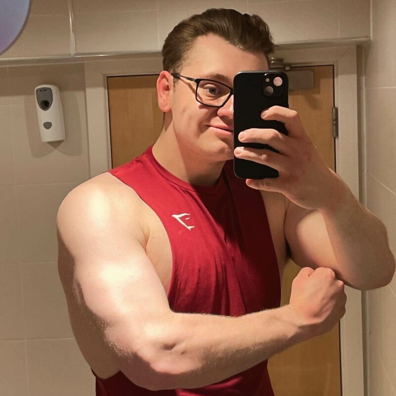 Man standing in front of the mirror wearing a red vest holding his phone in one hand and flexing his other arm