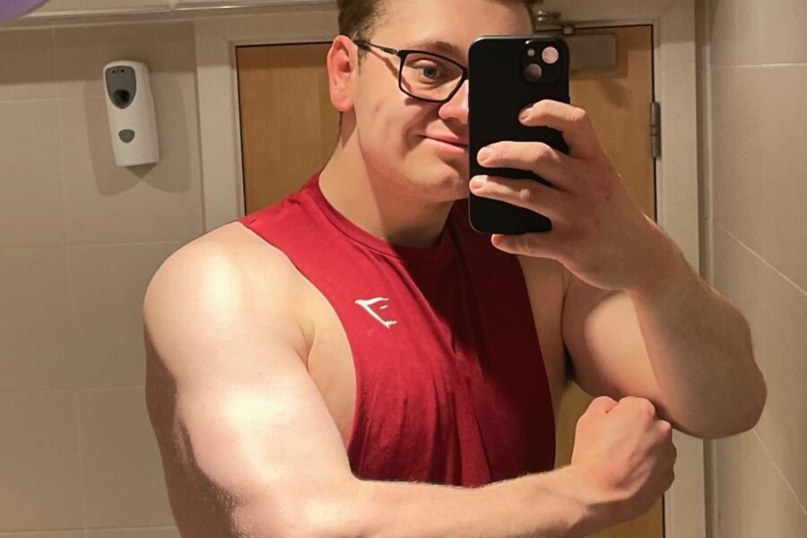 Man standing in front of the mirror wearing a red vest holding his phone in one hand and flexing his other arm