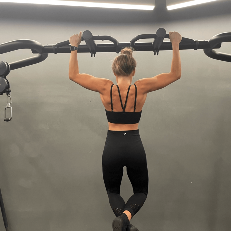 A woman facing away from the camera handing by her arms in a pull up position from gym equipment