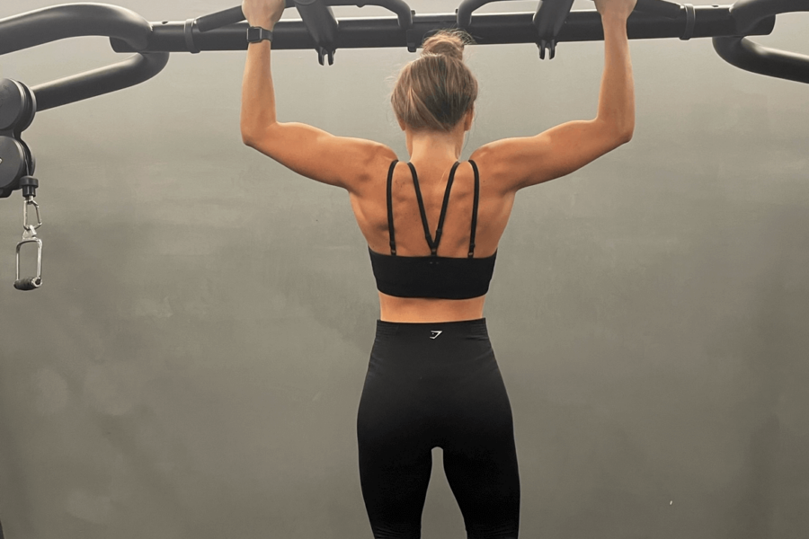 A woman facing away from the camera handing by her arms in a pull up position from gym equipment