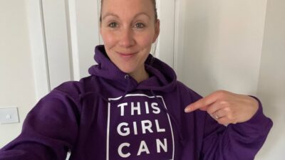 A woman with blonde hair facing the camera, pointing at her purpose This Girl Can jumper