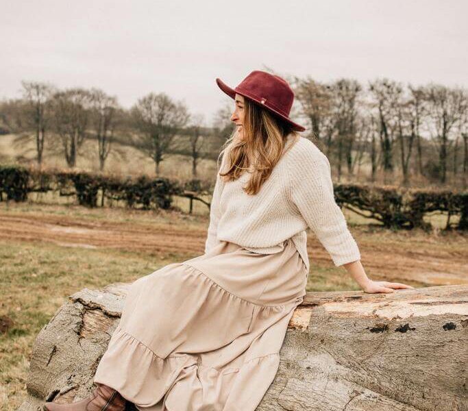 A woman facing away from the camera sitting on a felled tree wearing a hat