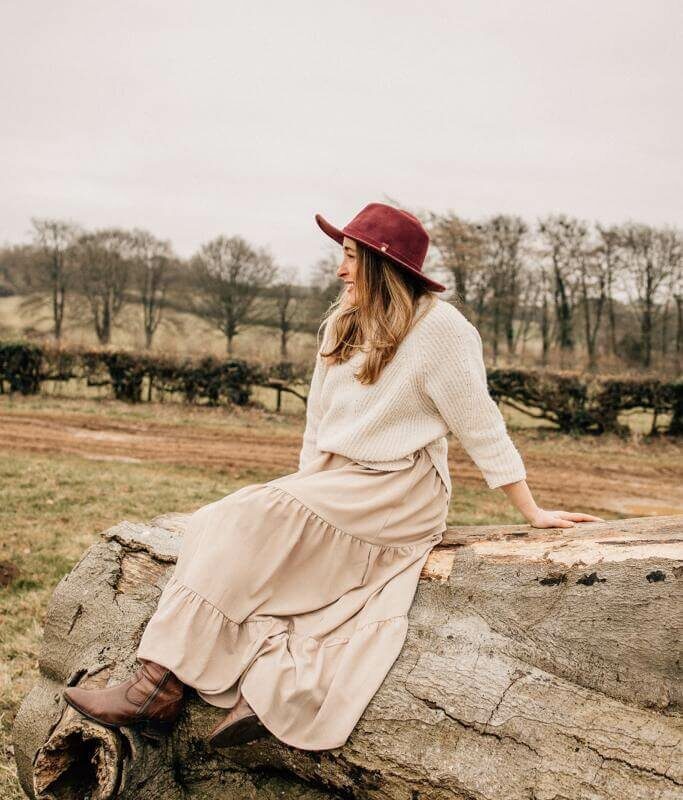 A woman facing away from the camera sitting on a felled tree wearing a hat