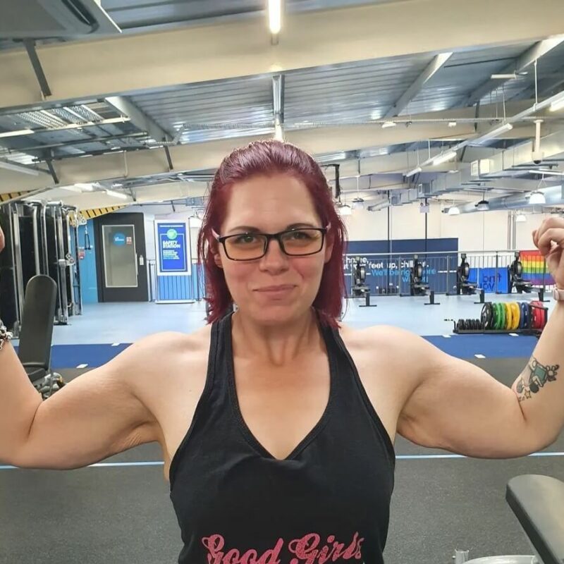A woman in glasses and a vest flexing her arms, facing the camera